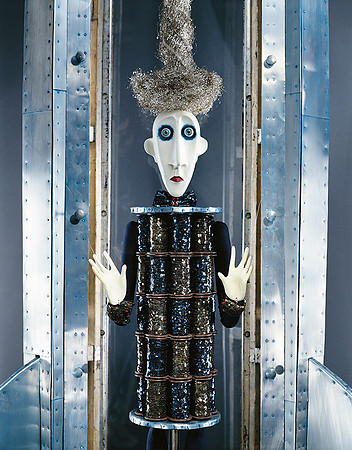 Fritz puppet body of brown sequence spools standing between two silver metal columns.